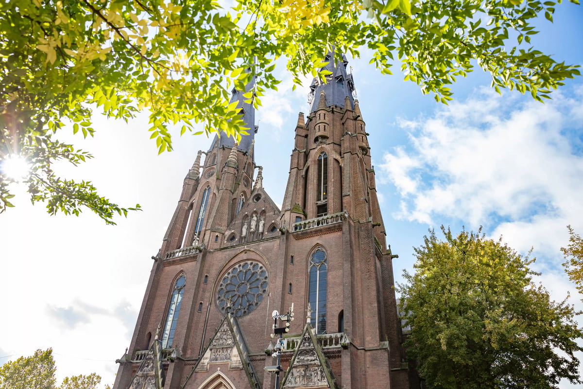 Église gothique en brique rouge avec deux hautes tours, encadrée par des feuilles vertes sous un ciel bleu