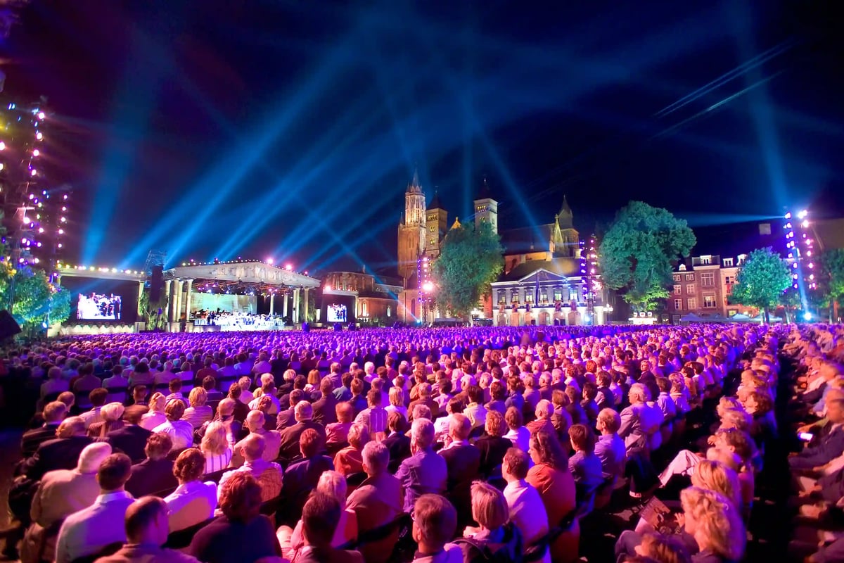 Grand concert en plein air de nuit avec scène illuminée, foule nombreuse et bâtiments historiques en arrière plan