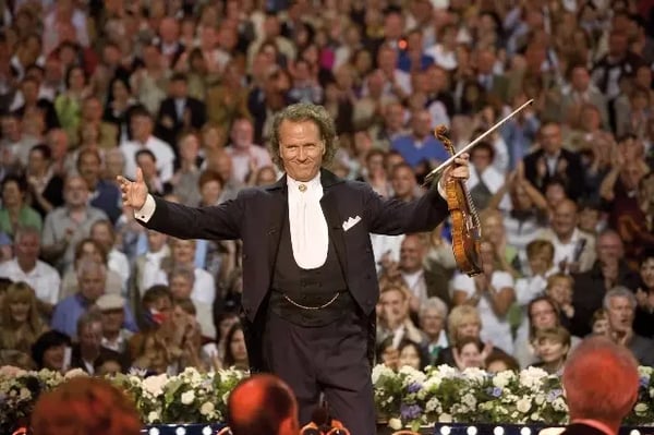 André Rieu steht auf der Bühne mit Geige und breitet die Arme vor einem grossem Publikum aus