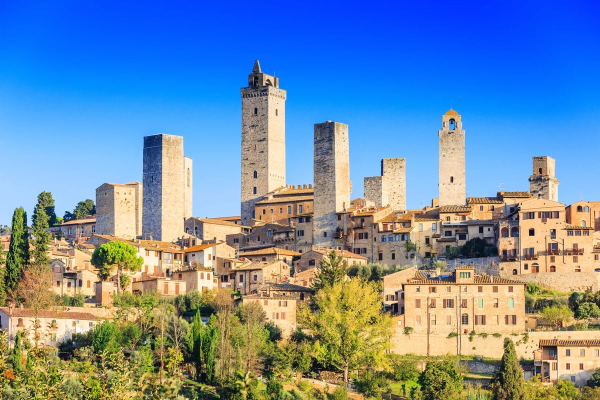 Vieille ville médiévale de San Gimignano avec plusieurs hautes tours en pierre au-dessus des toits en tuiles, sous un ciel bleu intense.