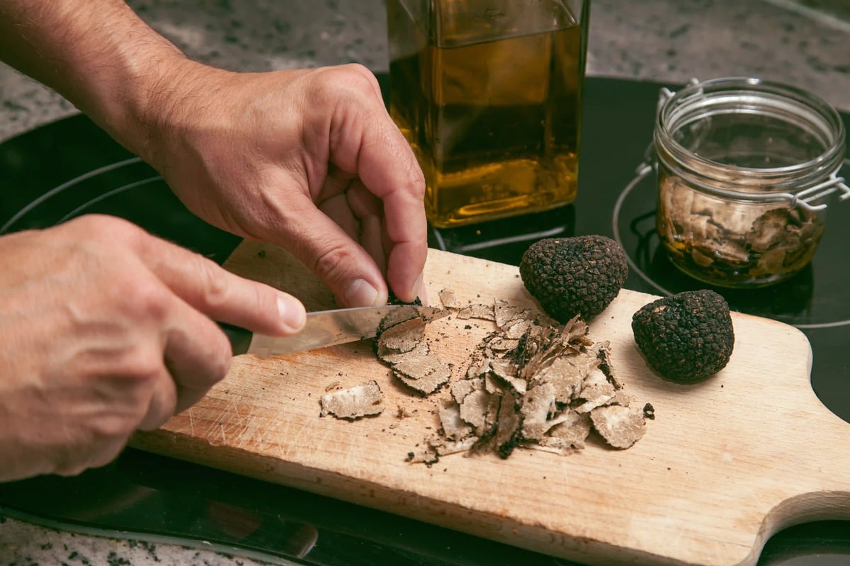 Gros plan: des mains tranchent une truffe en fines lamelles sur une planche en bois; à côté, des truffes entières, une bouteille d huile et un bocal.