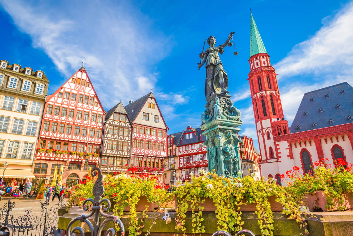 Der Frankfurter Römerberg mit dem Gerechtigkeitsbrunnen im Vordergrund und den charakteristischen rekonstruierten Fachwerkhäusern unter blauem Himmel