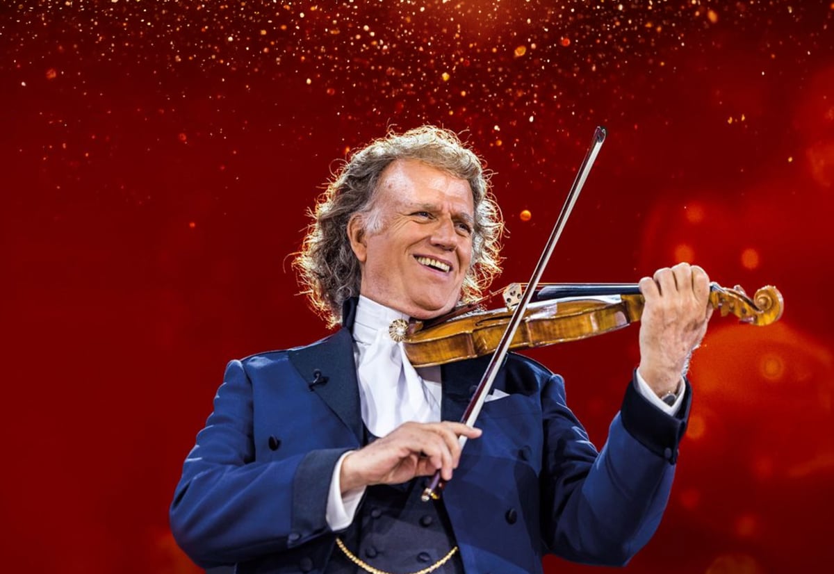 Porträt von André Rieu im blauen Frack, der lächelnd Geige spielt vor einem festlichen, rot-gold funkelnden Hintergrund.