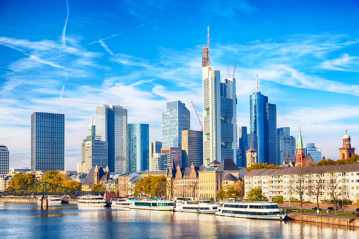 Die Frankfurter Skyline bei Tag mit Blick über den Main. Am Flussufer liegen mehrere Passagierschiffe vor den modernen Wolkenkratzern unter leicht bewölktem Himmel.