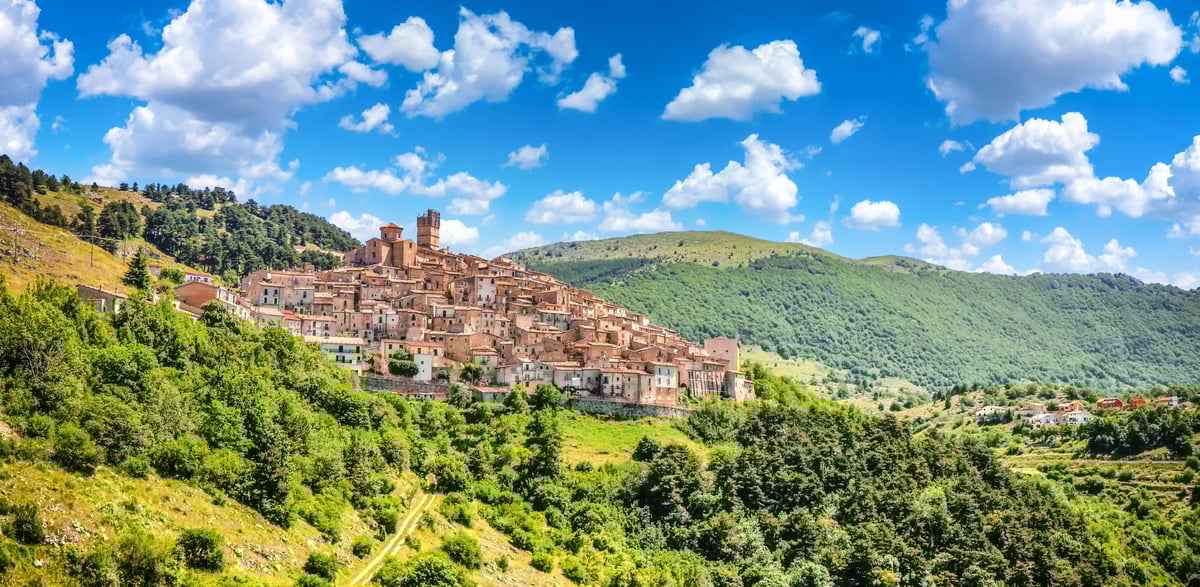 Mittelalterliche Altstadt von L’Aquila auf einem Hügel, umgeben von grünen Wäldern und Bergen unter blauem Himmel mit Wolken