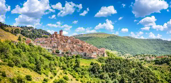 Mittelalterliche Altstadt von L’Aquila auf einem Hügel, umgeben von grünen Wäldern und Bergen unter blauem Himmel mit Wolken