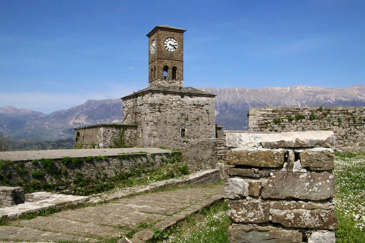 Forteresse en pierre avec tour horloge et remparts, chemin pavé au premier plan, chaîne de montagnes sous un ciel clair