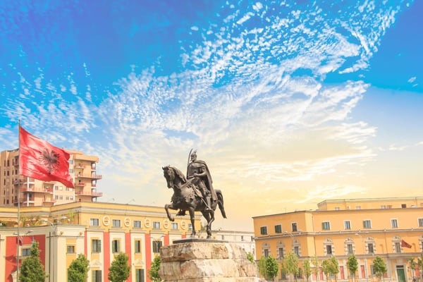 Reiterstatue auf Sockel auf einem Platz in Tirana, daneben weht eine rote Flagge, helle Gebäude und dramatische Wolken am Himmel