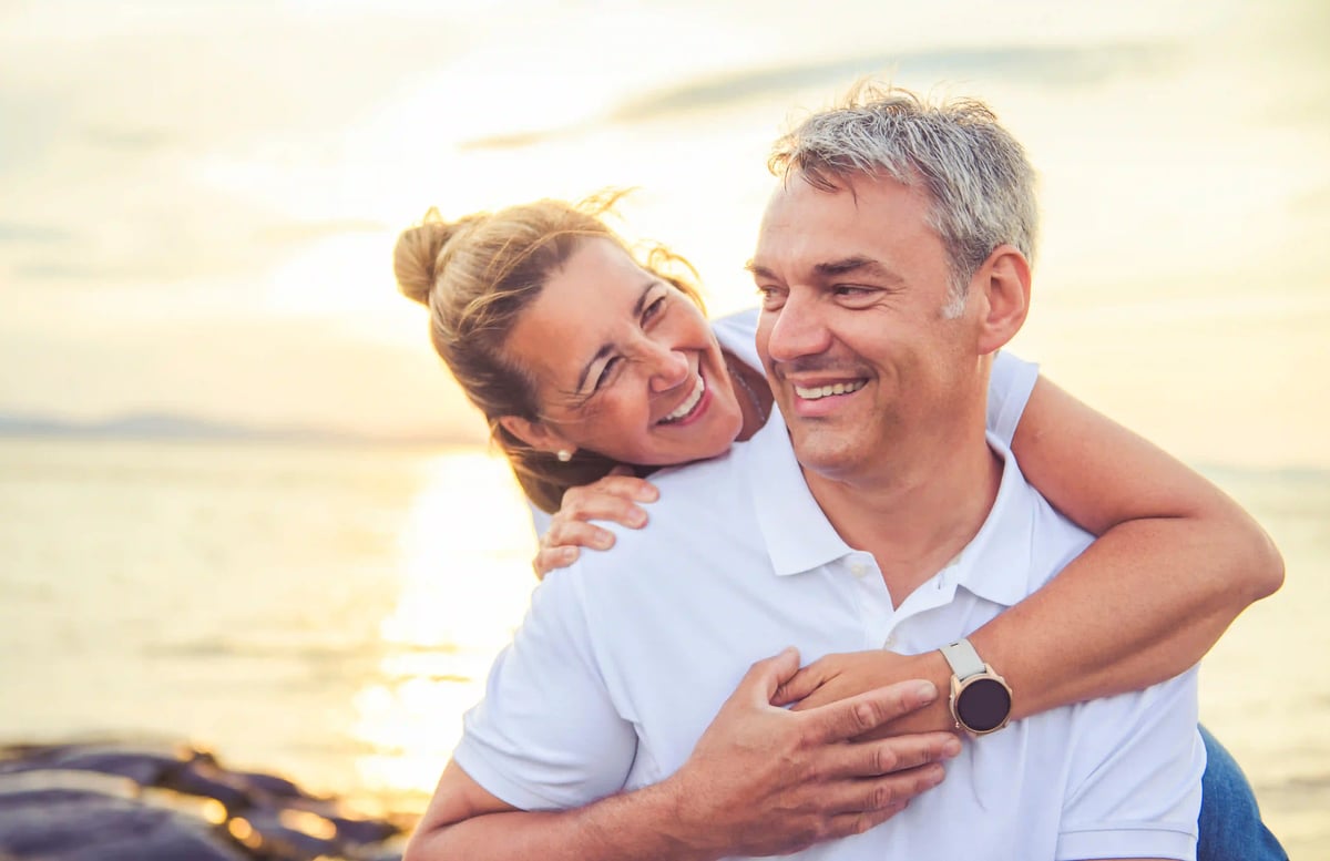 Couple souriant sur la plage au coucher du soleil, la femme enlace l homme par derrière, lumière chaude et mer en arrière-plan