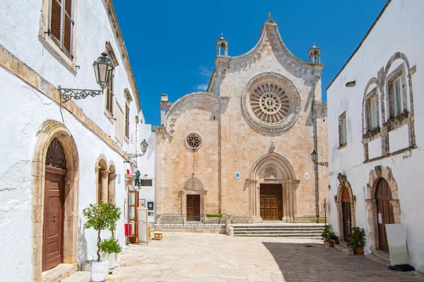 Helle Altstadtgasse in Ostuni mit historischer Kirche mit Rosettenfenster und umliegenden weissen Gebäuden unter blauem Himmel