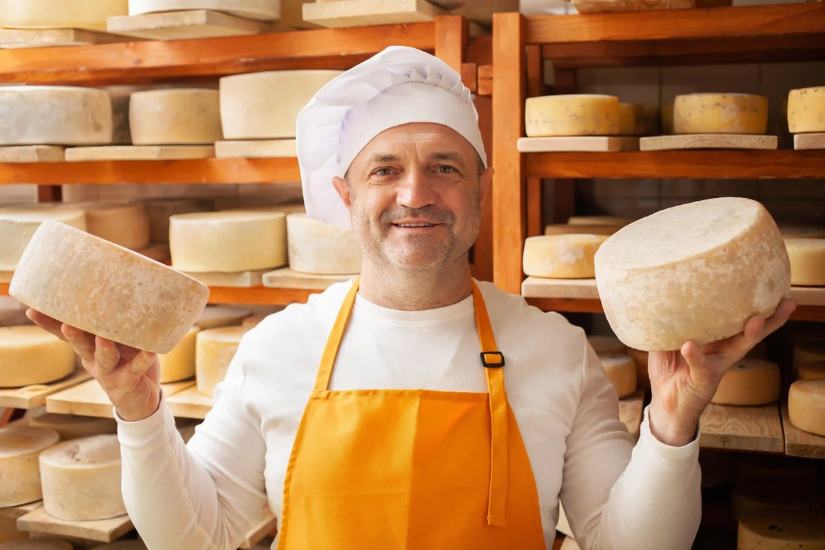 Käser in Schaukäserei hält zwei Laibe Käse vor Holzregalen mit gereiftem Käse
