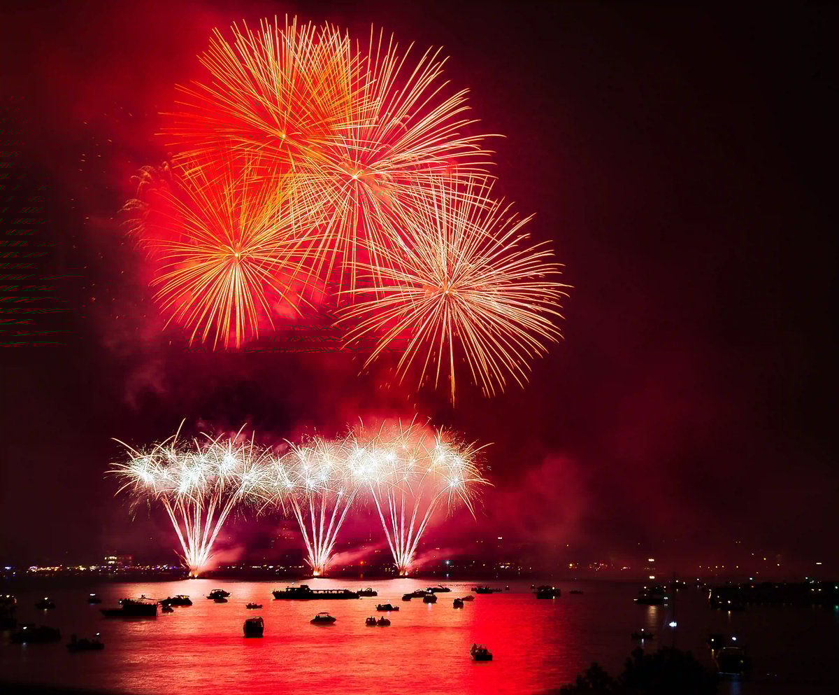 Rotes und goldenes Feuerwerk erleuchtet den Nachthimmel über einem See, die Lichtreflexionen spiegeln sich im ruhigen Wasser, mehrere Boote liegen im Vordergrund