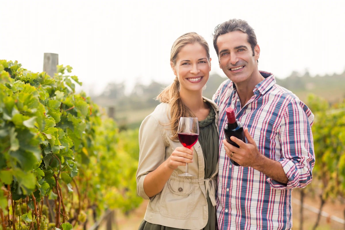 Lächelndes Paar steht im Weinberg und hält ein Glas Rotwein sowie eine Weinflasche in der Hand