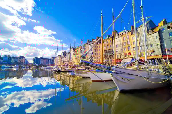 Historischer Hafen von Honfleur mit Segelbooten im Wasser, bunten Häuserfassaden und Spiegelungen unter blauem Himmel