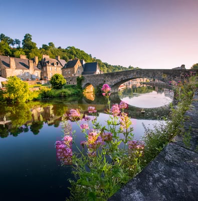 Malerische Flusslandschaft in Dinan mit steinerner Bogenbrücke, historischen Häusern, Spiegelungen im Wasser und blühenden Blumen im Vordergrund