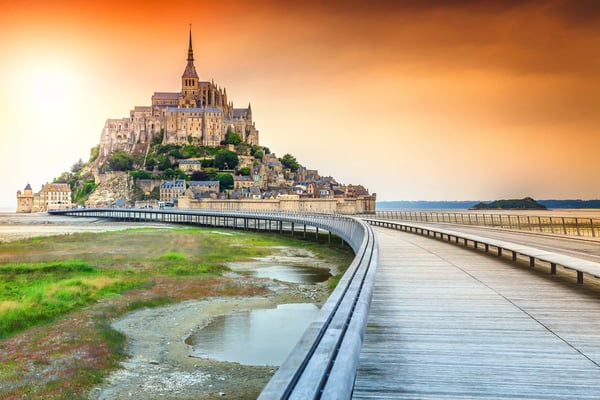 Mont Saint Michel mit Abtei auf einem Felsen, umgeben von Wattlandschaft und Holzsteg unter warmem Abendhimmel