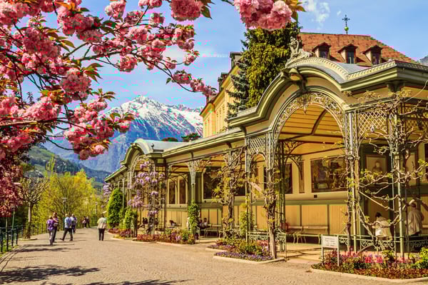 Elegante Wandelhalle in Meran mit blütenden Bäumen, gepflegten Blumenbeeten und Bergkulisse