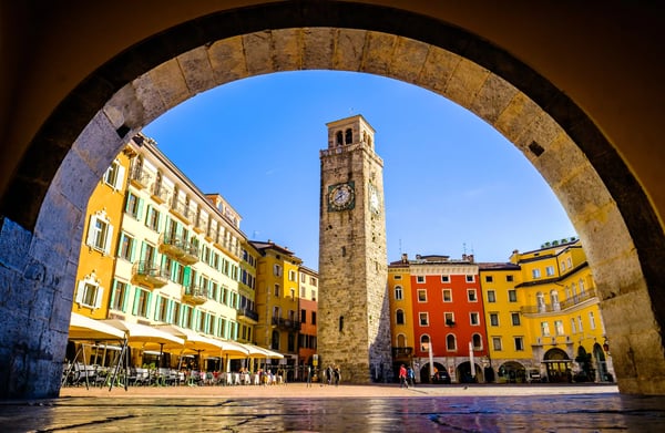 Blick durch einen steinernen Bogen auf einen sonnigen Platz mit historischem Uhrturm und farbigen Häuserfassaden