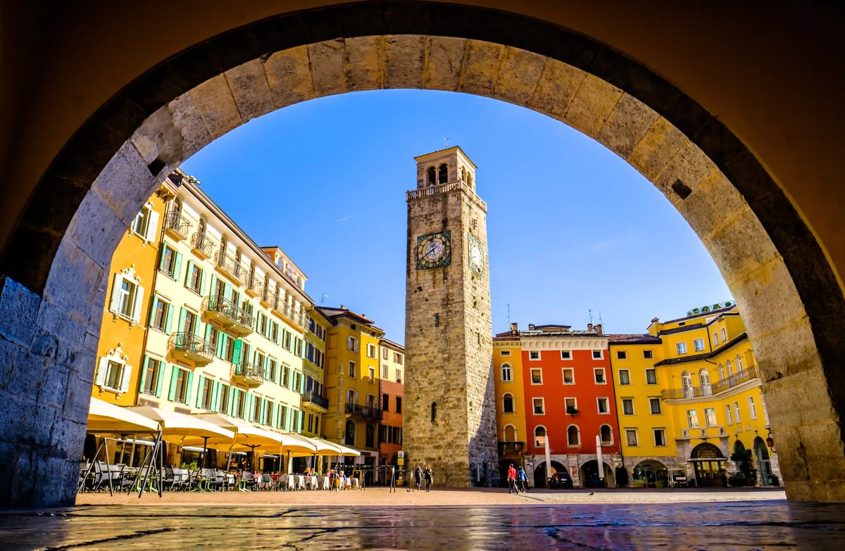 Vue a travers une arche en pierre sur une place ensoleillee avec une tour horloge historique et des facades colorees