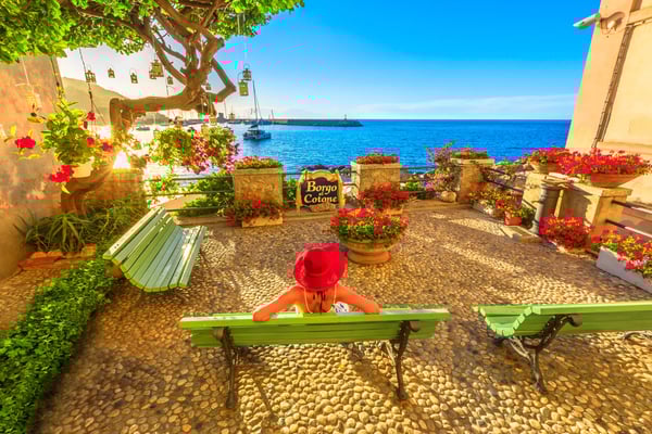 Sonnige Terrasse am Meer mit grünen Bänken, vielen pinken Blumen und einer Person mit rotem Hut, die auf das Wasser blickt.
