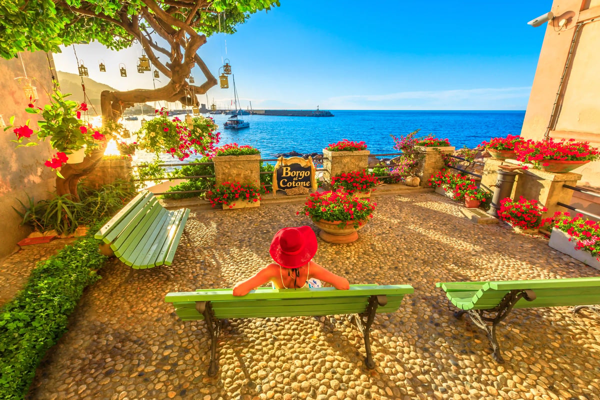 Sonnige Terrasse am Meer mit grünen Bänken, vielen pinken Blumen und einer Person mit rotem Hut, die auf das Wasser blickt.