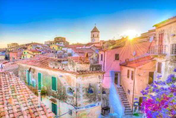 Pastellfarbene Häuser und rote Ziegeldächer im warmen Abendlicht, Kirchturm im Zentrum und Sonne tief am Himmel.