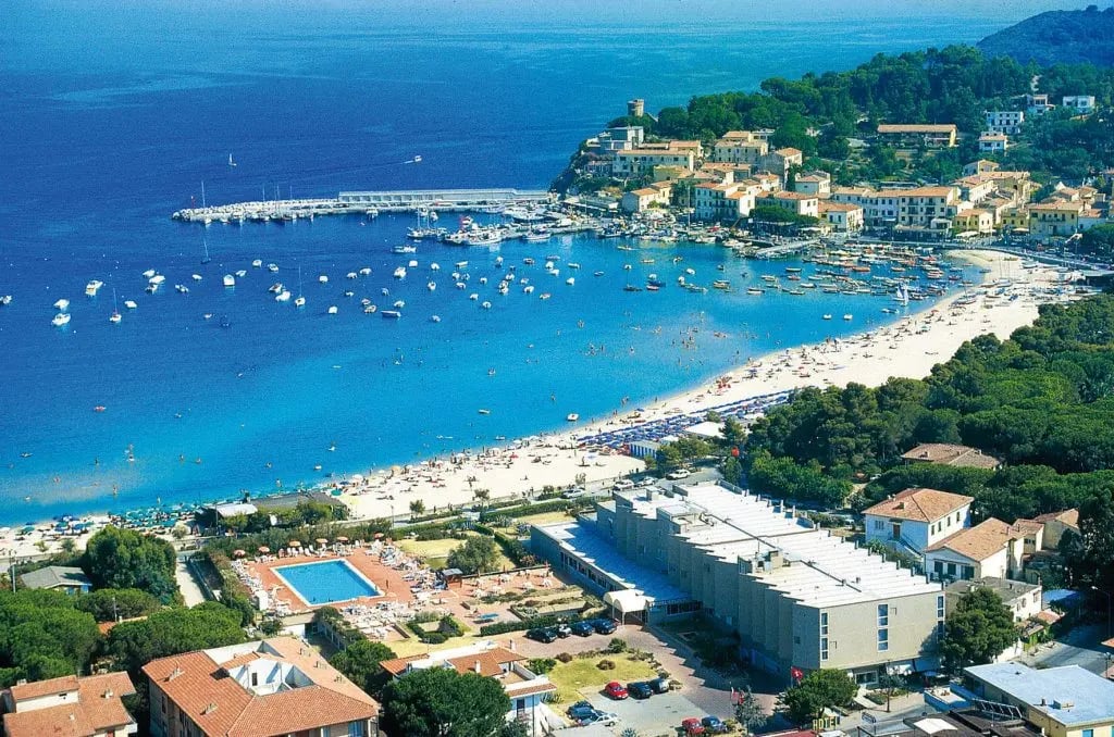 Blick auf Badebucht mit Sandstrand, zahlreichen Booten im türkisfarbenen Meer, Hafenanlage und Küstenort in hügeliger Landschaft.