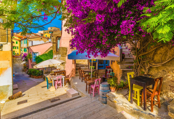 Kleine Altstadtgasse mit bunten Stühlen und Tischen unter Sonnenschirmen, überragt von üppiger, violetter Bougainvillea.