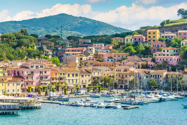 Blick auf Porto Azzurro mit bunten Häusern am Hafen, Segelbooten im türkisblauen Wasser und grünen Hügeln im Hintergrund.