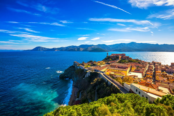 Panoramablick auf Portoferraio: Küstenstadt auf einer Landzunge, tiefblaues Meer, Berge am Horizont und Sonne über den Dächern.