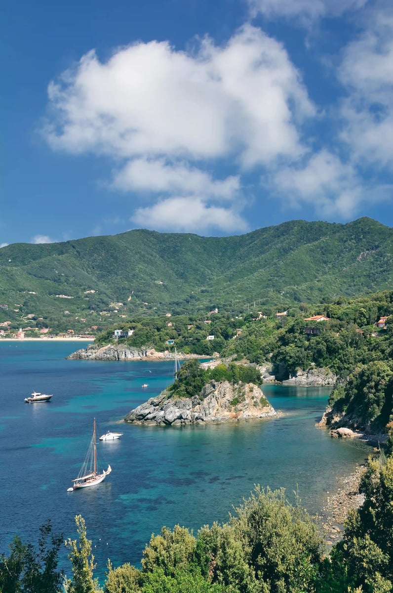 Ruhige Bucht mit klarem, blaugrünem Wasser, kleinen Segelbooten und bewaldeten Hügeln unter wolkigem Himmel
