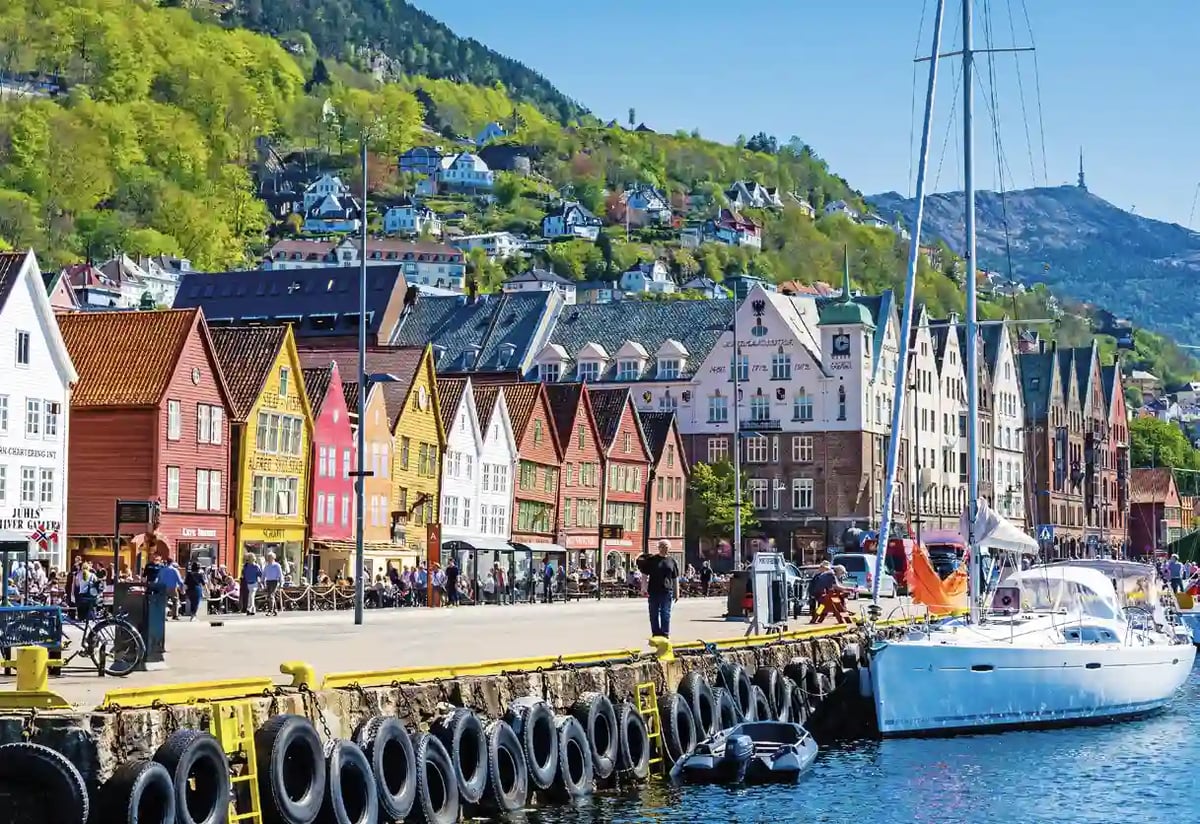 Reihe bunter Holzhäuser am Hafen von Bergen mit Booten im Vordergrund und bewaldetem Hang mit Wohnhäusern im Hintergrund