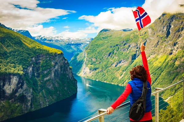 Person steht auf einer Aussichtsplattform über einem tief eingeschnittenen Fjord und hält eine norwegische Flagge hoch, steile grüne Felswände umgeben das Wasser