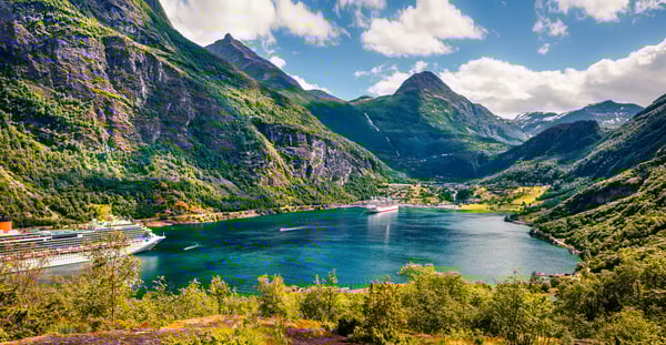Panoramablick über den Geirangerfjord mit steilen grünen Berghängen, einem Kreuzfahrtschiff im Fjord und verstreuten Höfen in der Landschaft