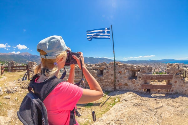 Reisende fotografiert eine weite Landschaft von einer historischen Ruine mit griechischer Flagge