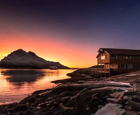 Holzhütte am felsigen Ufer bei Sonnenuntergang, ruhiges Wasser und Berge im Hintergrund