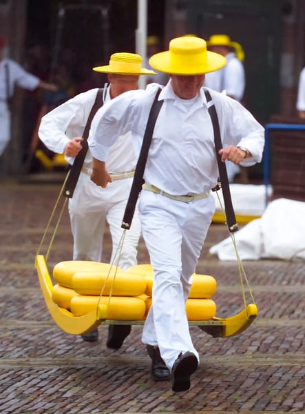 Käseträger in weisser Kleidung und gelben Hüten tragen Käse auf einer Holztrage über den Marktplatz