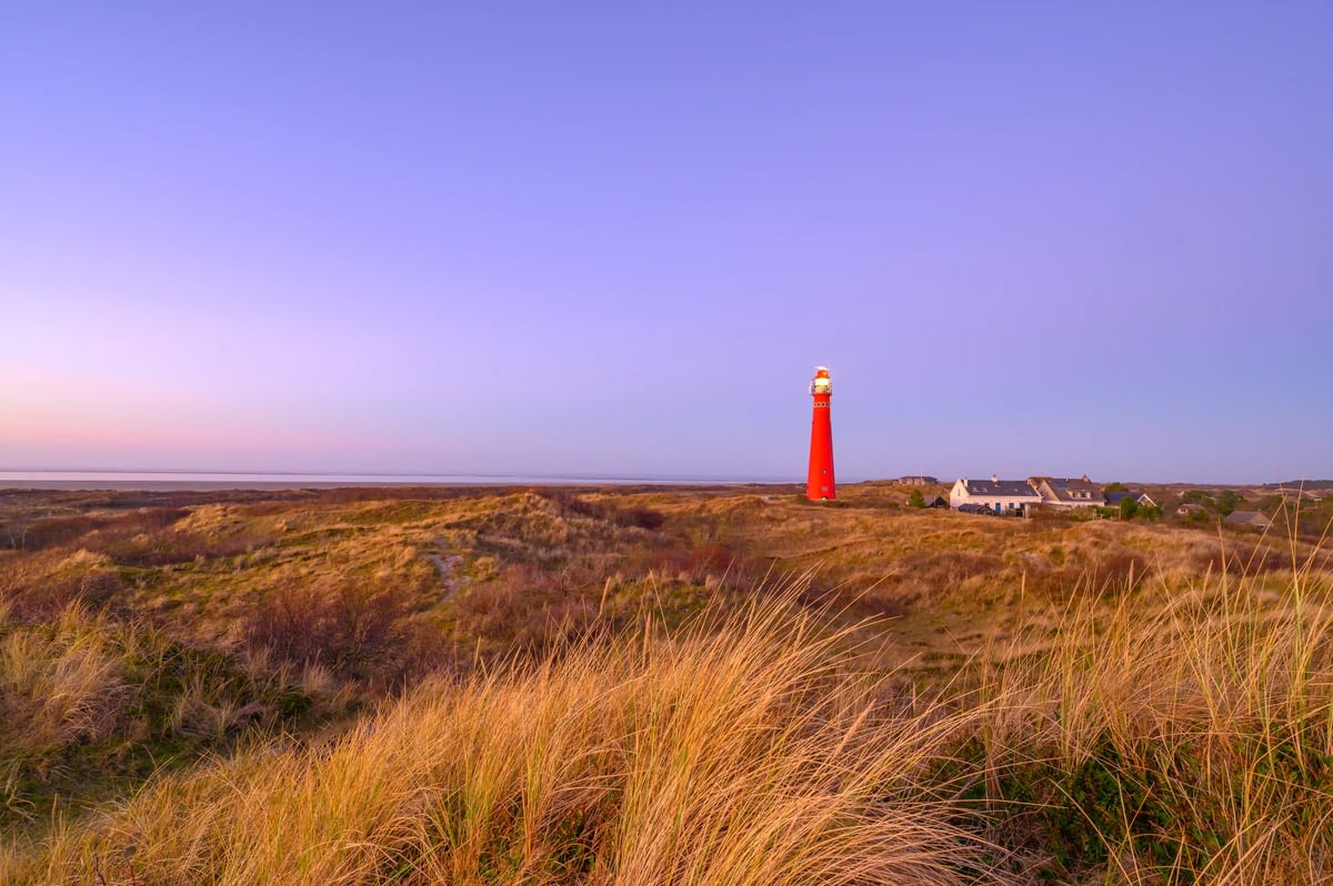 Roter Leuchtturm steht in einer Dünenlandschaft mit Gras und weiter Sicht bis zum Meer