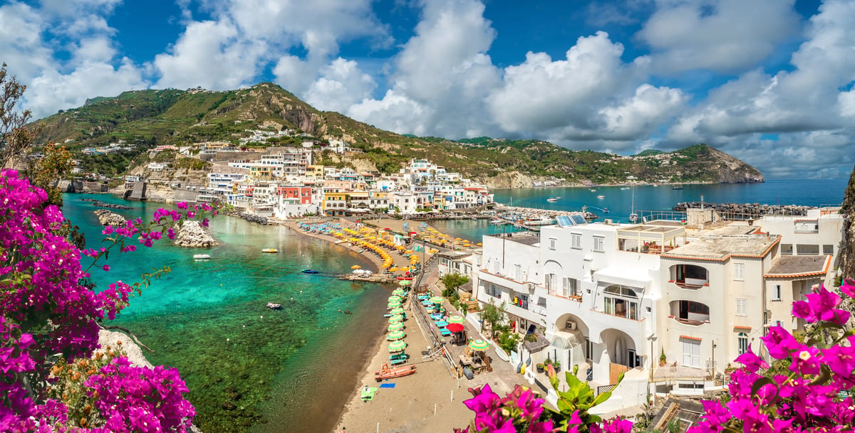 Blick über eine Bucht bei Sant Angelo auf Ischia mit Strandpromenade, bunten Sonnenschirmen, Booten und Hügeln am Meer.