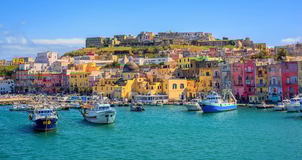Façades colorées du port de l île de Procida, nombreux bateaux sur une eau turquoise et forteresse sur la colline.