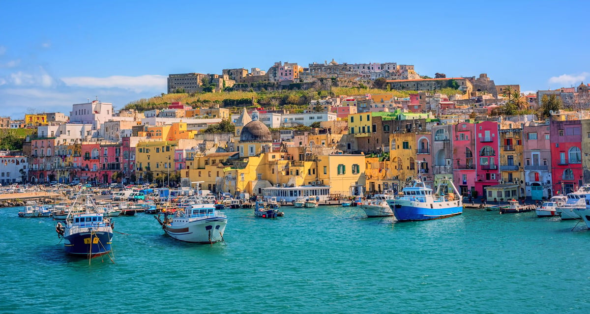 Bunte Häuserfronten am Hafen der Insel Procida, viele Boote im türkisfarbenen Wasser und Festung auf dem Hügel.