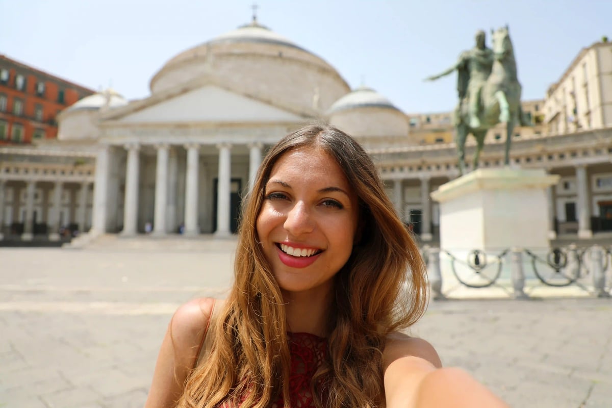 Frau macht ein Selfie auf der Piazza del Plebiscito in Neapel, im Hintergrund Säulenhalle und Reiterstatue bei Sonnenschein.