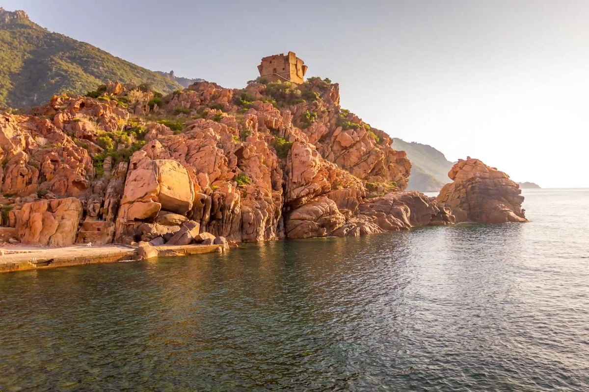 Rötliche Felsformationen der Calanches an der Küste mit kleinem Wachturm auf einem Felsen über ruhigem Wasser im Abendlicht