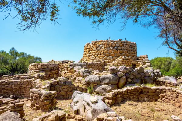 Steinruinen einer Nuraghe Anlage mit runder Turmstruktur und Trockenmauern in einer kargen Landschaft unter blauem Himmel