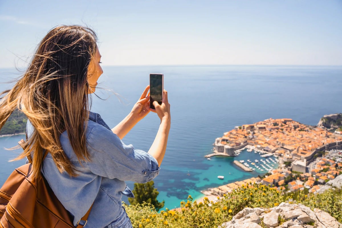 Frau mit Rucksack fotografiert mit dem Smartphone die Altstadt von Dubrovnik über dem tiefblauen Meer aus erhöhter Aussicht