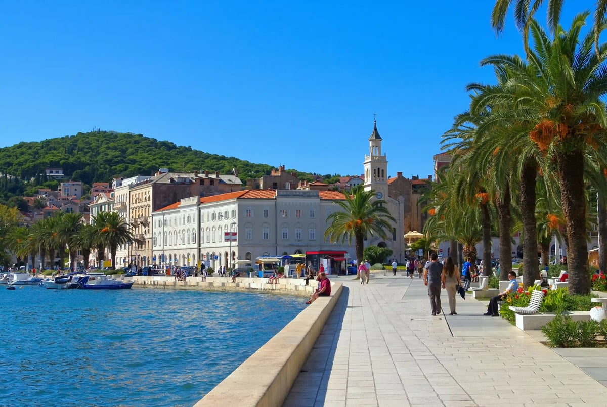 Promenade bordée de palmiers à Split avec port, bâtiments historiques et passants sous le soleil