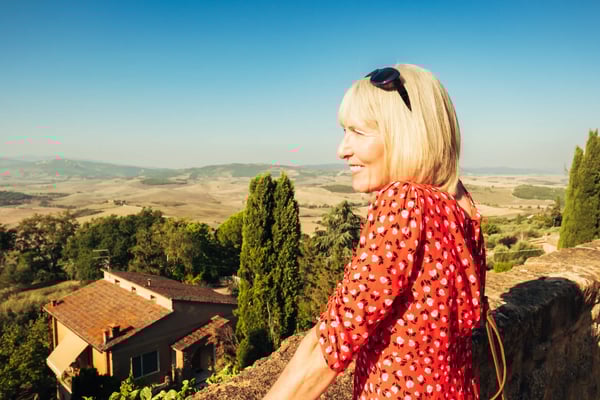 Frau in rotem Kleid steht an einer Mauer und blickt über eine weite Hügellandschaft mit Dörfern und Feldern