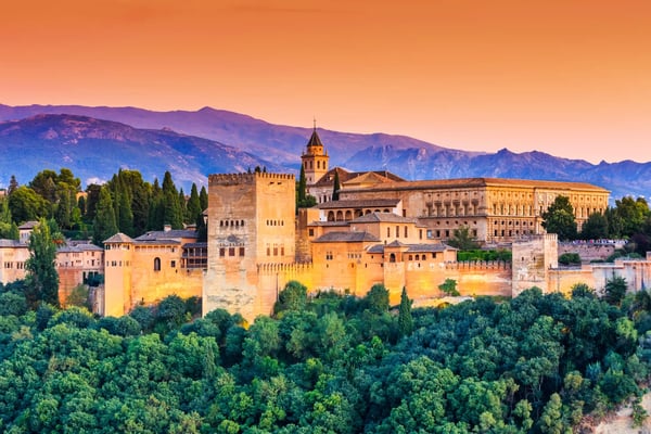 Alhambra in Granada bei Abenddämmerung, beleuchtete Mauern und Türme über grünen Bäumen vor Bergen.