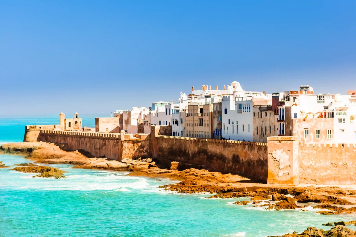 Stadtmauer und helle Häuser von Essaouira an der Atlantikküste, Brandung an felsigem Ufer unter blauem Himmel.