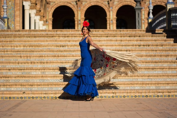 Flamenco-Tänzerin in blauem Kleid mit roter Blume, pose auf breiter Treppe vor Arkaden.